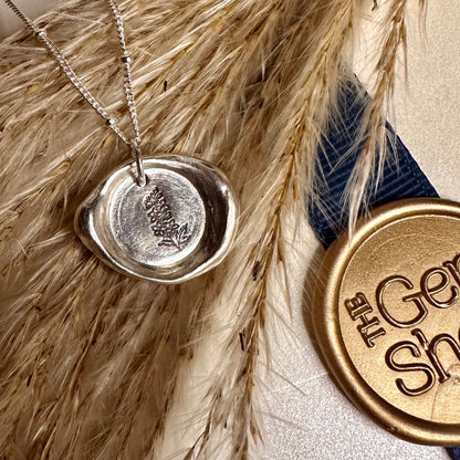 Silver necklace with a wax seal pendant on pampas grass, next to a gold 'The Gem Shed' logo.