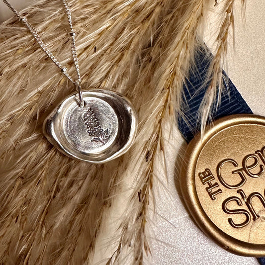 Silver necklace with a wax seal pendant on pampas grass, next to a gold 'The Gem Shed' logo.