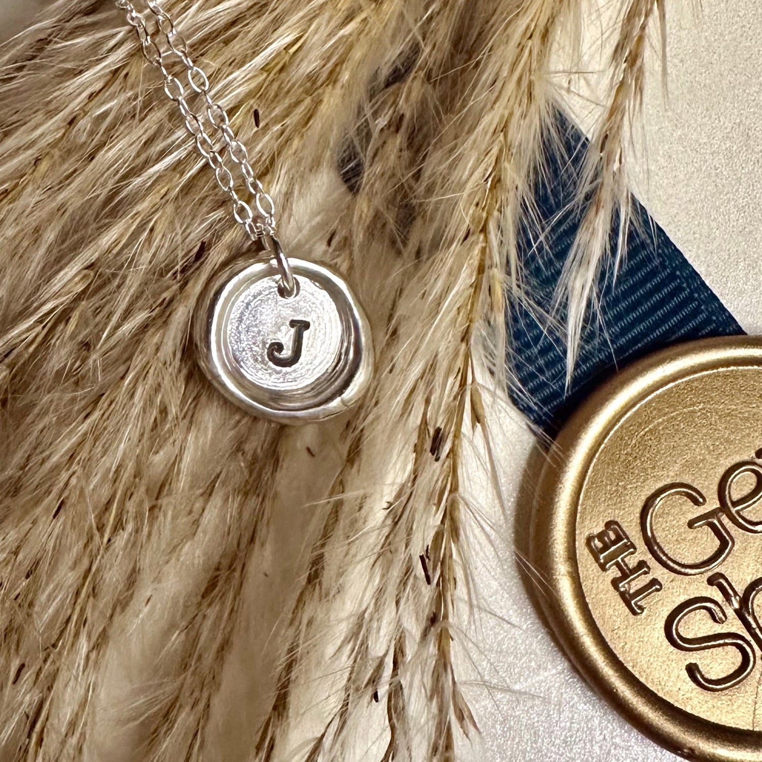 Silver necklace with a wax seal pendant featuring the letter 'J', placed on dried grass with a gold stamp and blue ribbon.